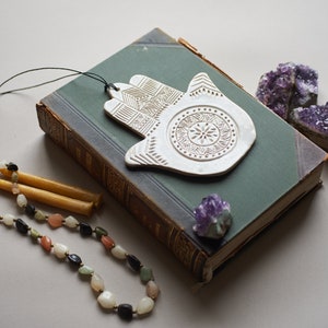 May include: White ceramic hamsa hand with intricate details, hanging from a black string. The hamsa is on top of a green book with a purple crystal next to it. A beaded necklace and two yellow candles are in the foreground.