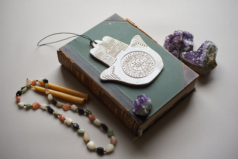 May include: White ceramic hamsa hand with intricate details, hanging from a black string. The hamsa is on top of a green book with a purple crystal next to it. A beaded necklace and two yellow candles are in the foreground.