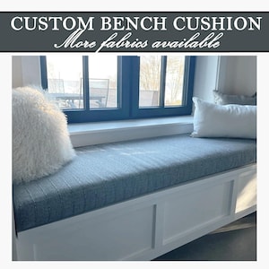 May include: A custom bench cushion with a gray fabric, white trim, and two decorative pillows. The text "CUSTOM BENCH CUSHION" is at the top of the image, with "More fabrics available" below it.