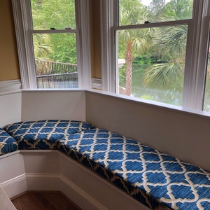 May include: A white window seat with a blue and gold patterned cushion. The seat is in a corner room with a window overlooking a lake and palm trees.