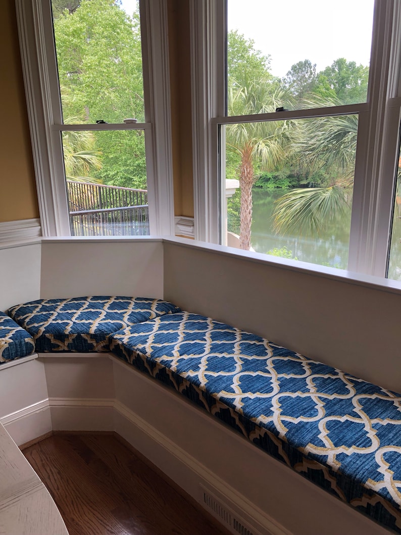 May include: A white window seat with a blue and gold patterned cushion. The seat is in a corner room with a window overlooking a lake and palm trees.