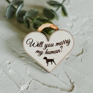 May include: A heart-shaped wooden sign with the text "Will you marry my human?" in elegant script. A small silhouette of a dog is below the text. The sign has a light wood grain and a small loop at the top.