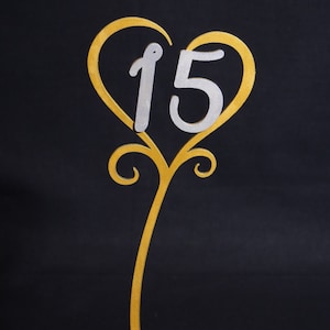 May include: A gold-colored table number stand with a heart-shaped top. The number "15" is in silver. The stand has a curved stem and a base with decorative swirls. The background is black.