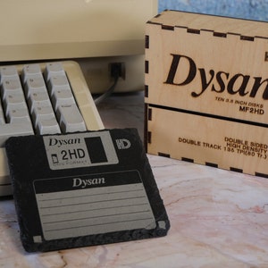 May include: A black Dysan 2HD 3.5-inch floppy disk next to a wooden box with the Dysan logo and text. The box is labeled "TEN 3.5 INCH DISKS MF2HD". A vintage computer keyboard is in the background.