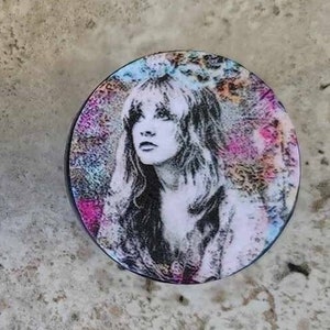 Può includere: Due sottobicchieri rotondi e colorati con un'immagine in bianco e nero di una donna con i capelli lunghi. I sottobicchieri hanno uno sfondo testurizzato, astratto con sfumature di rosa, blu e oro.