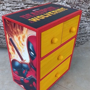 May include: A wooden box with four drawers painted yellow. The box is decorated with a comic book image of Deadpool and Wolverine. The text on the box reads "Deadpool & Wolverine".