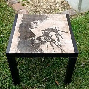 May include: Black and white image of a person with long hair and a long, sharp object in their hand. The image is printed on a wooden table with a black frame.