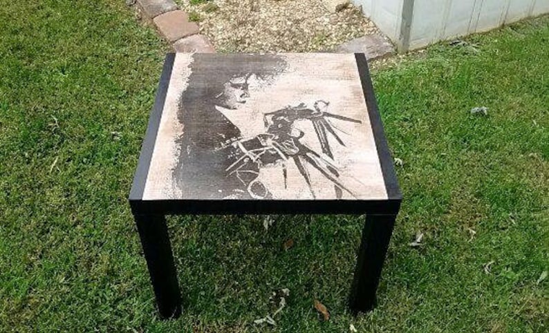 May include: Black and white image of a person with long hair and a long, sharp object in their hand. The image is printed on a wooden table with a black frame.