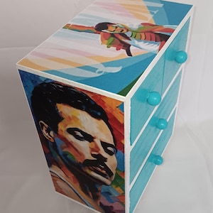May include: A small, three-drawer wooden chest with a teal finish and teal knobs. The front features a colorful portrait of a man with a mustache. The top has a colorful illustration of a person in flight.