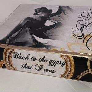 May include: A wooden box with a black and gold design. The top of the box features a black and white photo of a woman wearing a hat and a flowing dress. The text "Back to the gypsy that I was" is printed on the front of the box. The text "Stevie Nicks" is printed on the side of the box.