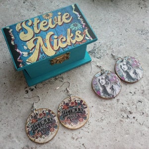 May include: A set of four earrings featuring Stevie Nicks and Fleetwood Mac designs. The earrings are made of wood and have silver hooks. The earrings are packaged in a blue wooden box with the words "Stevie Nicks" printed on the top.