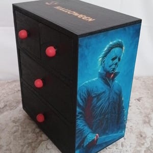May include: A black wooden storage box with three drawers and red knobs. The side of the box features a portrait of a figure in a blue jacket holding a knife. The word "HALLOWEEN" is printed on the top of the box.