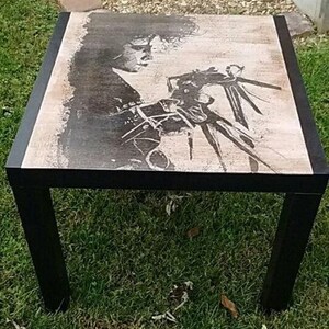 May include: Black coffee table with a printed image of a person with long hair and a creature with long arms and claws. The table has a black frame and a light brown top.