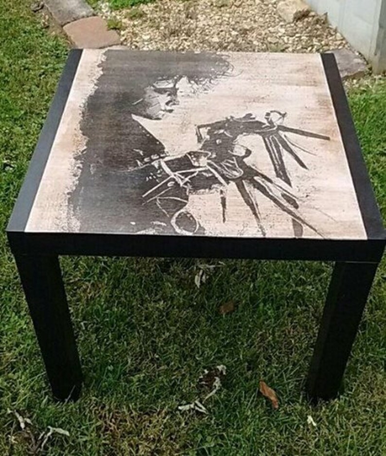 May include: Black coffee table with a printed image of a person with long hair and a creature with long arms and claws. The table has a black frame and a light brown top.