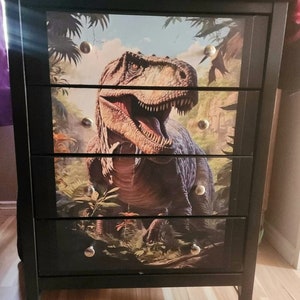 Puede incluir: Cómoda negra con cuatro cajones con estampado de dinosaurio. Los cajones tienen pomos plateados. El dinosaurio es un Tyrannosaurus Rex con un esquema de color verde, marrón y amarillo. El dinosaurio está en un entorno de jungla con plantas y árboles verdes.