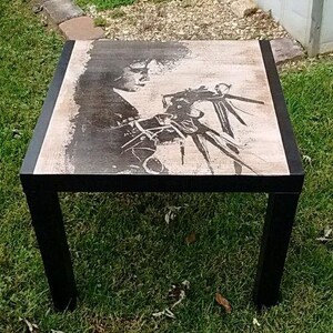 May include: Black coffee table with a square top featuring a black and white image of a person with a large hand. The table has a black frame.