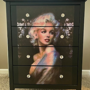 Puede incluir: Cómoda negra con tres cajones que presentan una imagen vintage de Marilyn Monroe. Cada cajón tiene una perilla de cristal.