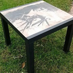 May include: A square black metal table with a white top featuring a black and white graphic print. The table is sitting on a green grassy surface.