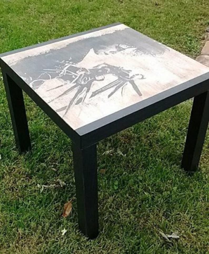 May include: A square black metal table with a white top featuring a black and white graphic print. The table is sitting on a green grassy surface.