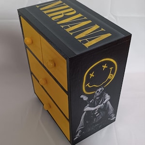 Puede incluir: Una caja de madera negra con cuatro cajones amarillos y un logotipo de Nirvana en la parte superior. La parte delantera de la caja presenta una imagen en blanco y negro de una persona con una guitarra y el icónico logotipo de la cara sonriente de la banda.