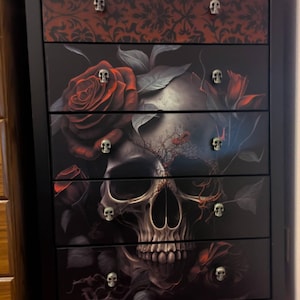 Könnte beinhalten: Schwarze Kommode mit sechs Schubladen, mit einem Totenkopf- und Rosenmuster. Die oberste Schublade hat ein rotes Blumenmuster. Jede Schublade hat einen knauf in Schädelform. Der Stil ist detailliert und gotisch.