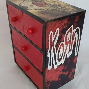 Puede incluir: Una caja de madera roja con cuatro cajones y un borde negro. La caja está decorada con un gráfico de la banda Korn y un patrón tie-dye. Los cajones tienen pomos rojos.