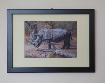 May include: Framed artwork featuring a detailed drawing of a rhinoceros in a natural setting. The rhino is depicted in shades of gray, with a prominent horn. The artwork is set against a beige background, framed in a black frame.
