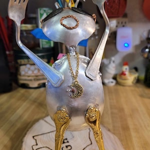 May include: A silver and gold sculpture of a person made from repurposed silverware. The sculpture is wearing sunglasses, a gold chain necklace with a circular pendant, and has its arms raised in the air. The sculpture is standing on a wooden base with the word "BURGER" carved into it.