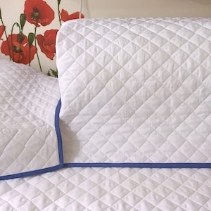 Peut inclure: Housse de machine à coudre matelassée blanche avec une bordure bleue. La housse est faite d'un tissu matelassé doux et présente un motif en losange.