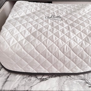 May include: A white quilted appliance cover with a diamond-stitched pattern and a dark gray bottom border. The word Bella is embroidered in black cursive script on the top center of the cover, featuring a decorative flourish underneath the text.
