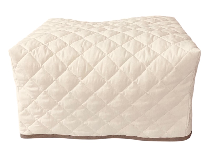 May include: A rectangular appliance cover in a light cream color featuring a quilted diamond pattern across the entire surface. The cover has a smooth, structured shape with a thin, dark brown trim along the bottom edge, providing a clean finish.
