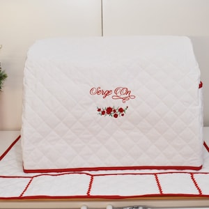 May include: White quilted sewing machine cover with red trim and a red embroidered design of roses and the text "Serge On".