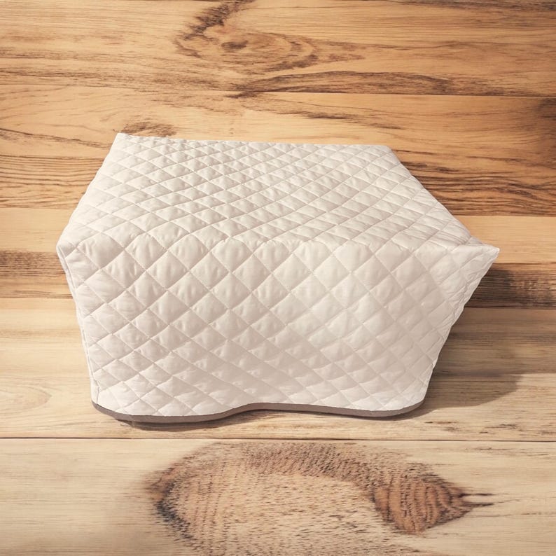 May include: A white quilted appliance cover featuring a diamond-shaped stitched pattern across the entire surface. The cover has a structured, rectangular shape with a smooth, finished bottom edge, designed to fit over kitchen equipment.