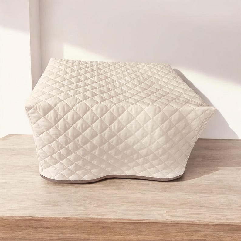 May include: A cream-colored, quilted appliance cover featuring a diamond-stitched pattern across its entire surface. The cover has a structured, rectangular shape with a flat top and slightly tapered sides, finished with a solid-colored bottom trim.