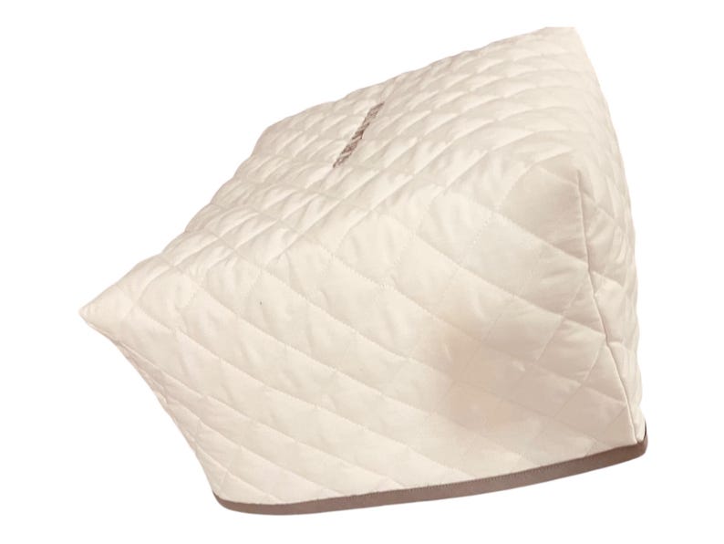 May include: A cream-colored, quilted appliance cover featuring a diamond-stitched pattern across its exterior. The cover has a structured, boxy shape with a solid, darker-toned trim along the bottom edge, designed to fit over a kitchen appliance.