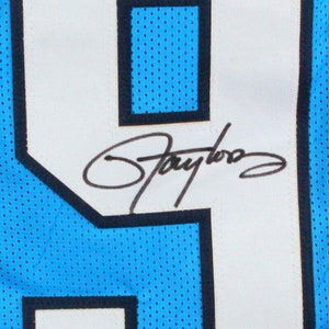 May include: Close-up of a light blue jersey with a large white number 9 outlined in black. The number has a black signature and a small authentication sticker at the bottom. The jersey has a mesh texture.