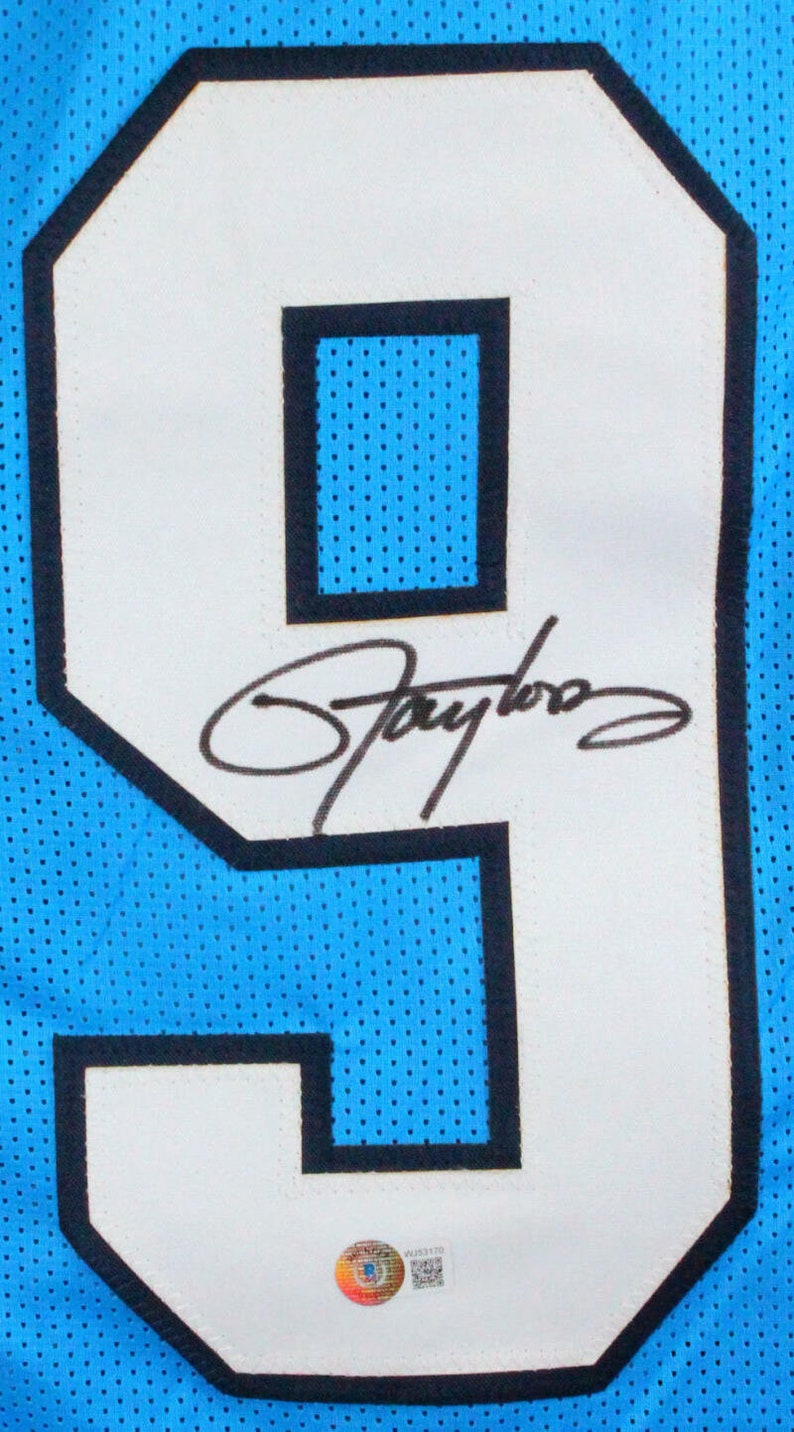 May include: Close-up of a light blue jersey with a large white number 9 outlined in black. The number has a black signature and a small authentication sticker at the bottom. The jersey has a mesh texture.