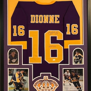 May include: A framed purple and gold hockey jersey with the name "DIONNE" and the number "16" on the back. The jersey is signed by the player and includes a small photo of the player in action. The jersey is framed with a purple mat and a black frame.
