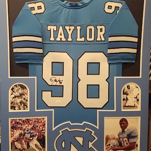 May include: Framed light blue football jersey with the name "TAYLOR" and the number "98". The jersey is displayed with photos and the team logo. The jersey is signed. The frame is light blue.