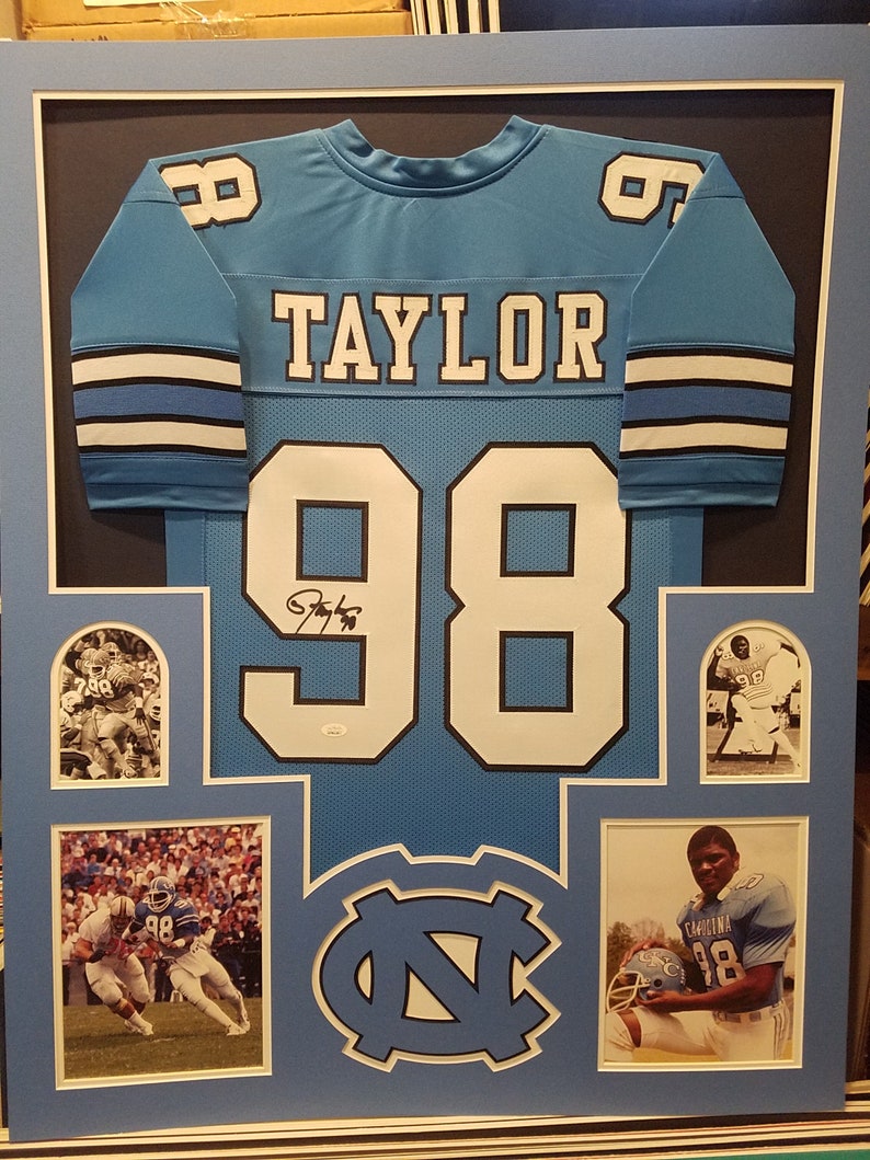 May include: Framed light blue football jersey with the name "TAYLOR" and the number "98". The jersey is displayed with photos and the team logo. The jersey is signed. The frame is light blue.