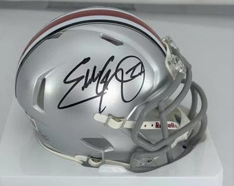 May include: A silver football helmet with a red and white stripe, signed with a black signature. The helmet has a face mask and a white logo on the side.