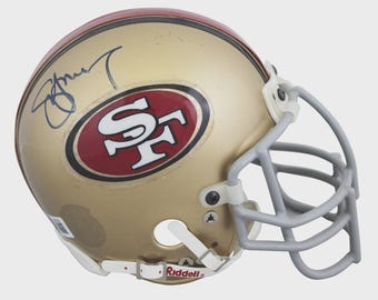 May include: A gold San Francisco 49ers football helmet featuring a red and white team logo on the side, a grey face mask, and a white chin strap with the Riddell brand name. A dark blue ink signature is visible on the upper left side of the helmet.