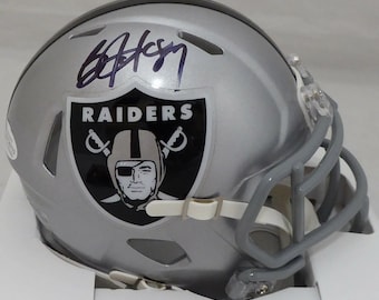 May include: A silver football helmet with a black and white Raiders logo and a purple signature. The helmet has a clear face mask.