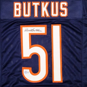 Puede incluir: Una camiseta de fútbol azul marino con el nombre "BUTKUS" y el número "51" en blanco con contornos naranja. La camiseta está firmada con tinta negra.