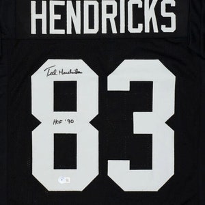 May include: Black football jersey with the name "HENDRICKS" and the number "83" in white. The jersey is signed "Ted Hendricks" and includes the text "HOF '90".