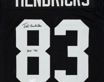 May include: Black football jersey with the name "HENDRICKS" and the number "83" in white. The jersey is signed "Ted Hendricks" and includes the text "HOF '90".