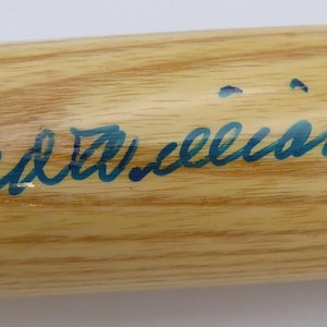 Pode incluir: Um bastão de beisebol de madeira com acabamento castanho claro. O bastão apresenta uma assinatura azul e uma pequena imagem na extremidade. O texto "S.A." é visível perto da pega.