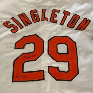May include: White baseball jersey with the name "SINGLETON" and the number "29" in orange with black outlines. The jersey is signed in black ink.