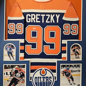 May include: A framed hockey jersey with the name "Gretzky" and the number "99" on the back. The jersey is orange, blue, and white with the Oilers logo on the front. The frame includes photos of Wayne Gretzky playing hockey.