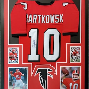 May include: A framed red Atlanta Falcons jersey with the name "Bartkowski" and the number "10" on the back. The jersey is signed by the player. The frame includes photos of the player in action.